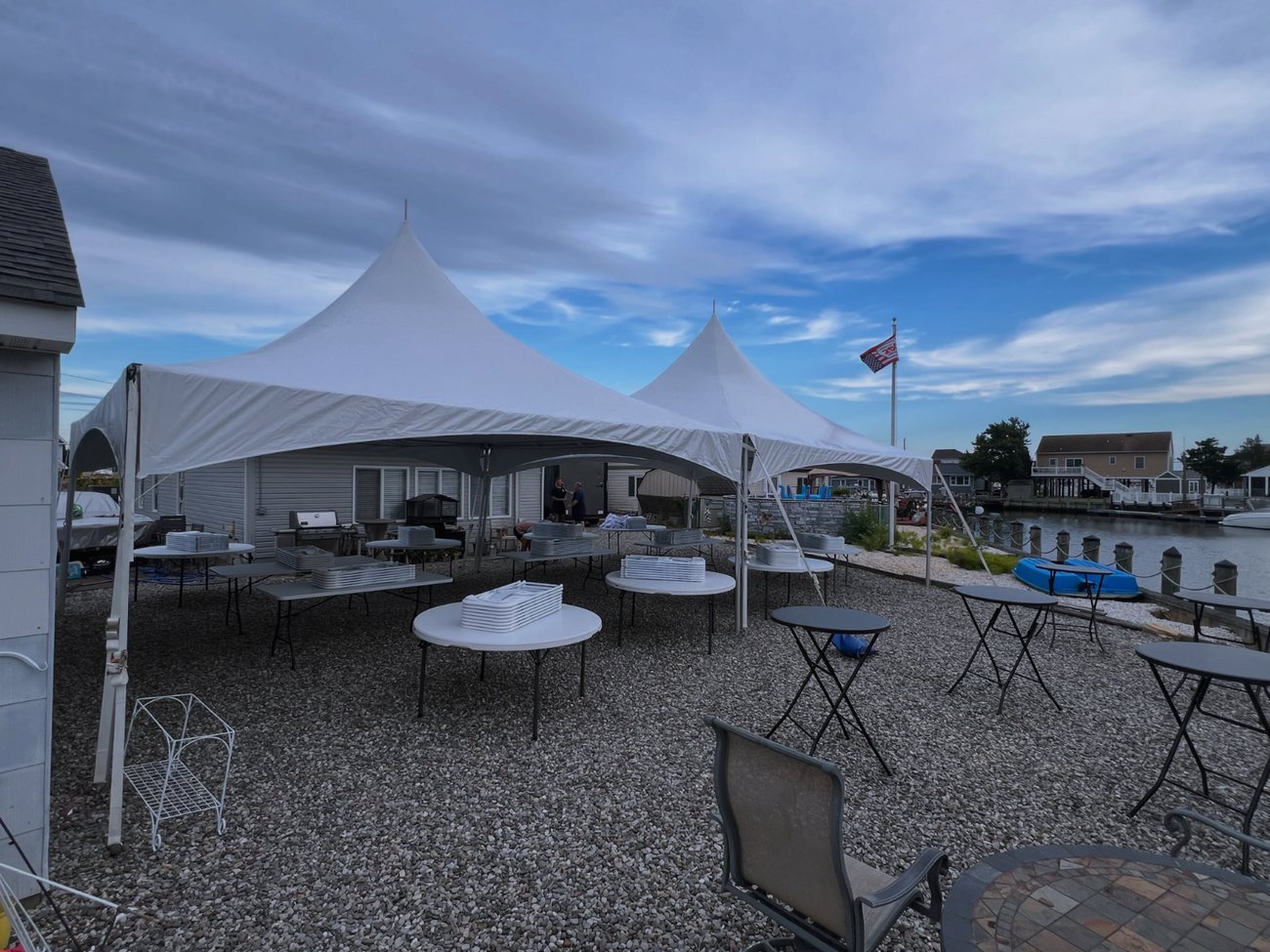 Two white high peak tents set up on a gravel bayfront yard with folding tables, a flag flying and boats at the dock behind.