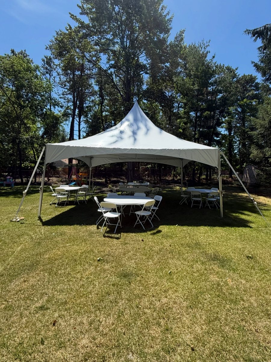 A 20 by 20 high peak tent in a pine-shaded backyard with a 60 inch round table and white folding chairs underneath.