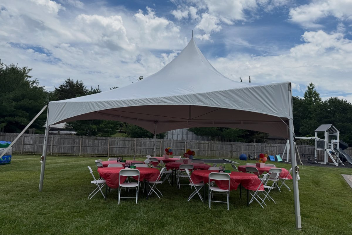 A 20 by 20 high peak tent set up on a fenced backyard lawn with round tables in red linens and white folding chairs.
