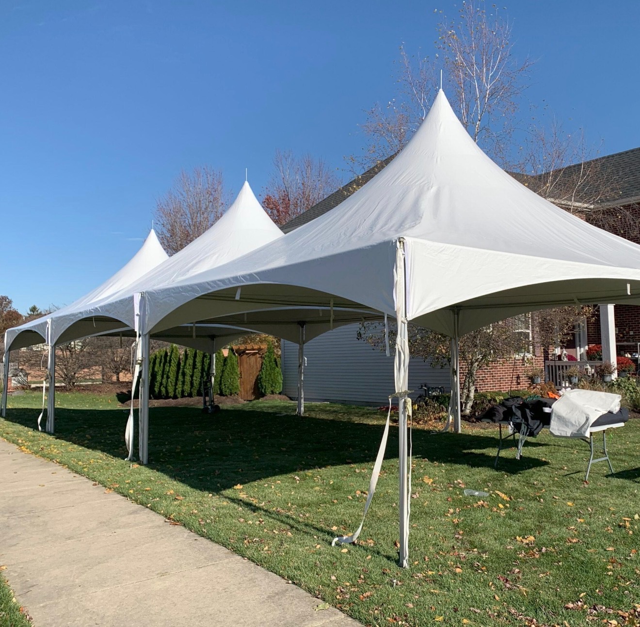 A 20 by 60 triple peak high peak tent set up on a front lawn beside a driveway under a clear blue sky.